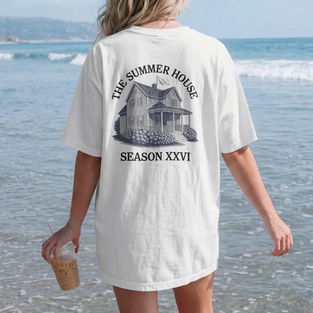 Woman on the beach in a White tee, back view: a shingled summer house with a porch and hydrangeas, under The Summer House and Season XXVI, as a fine engraving in navy and antique gold.