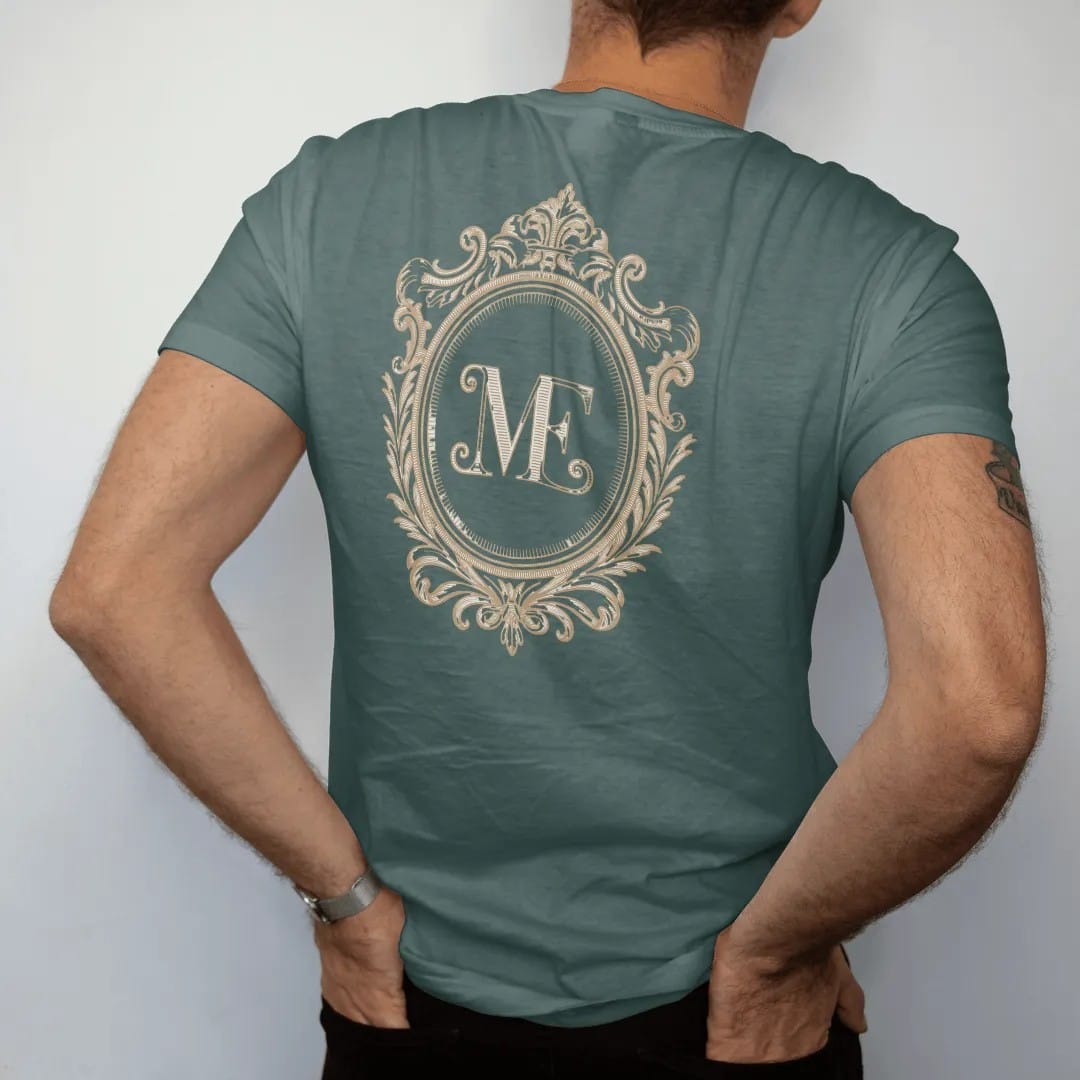 Man wearing a Blue Spruce tee, seen from the back: an ornate interlocking ME monogram, as a fine engraving in cream and champagne gold.