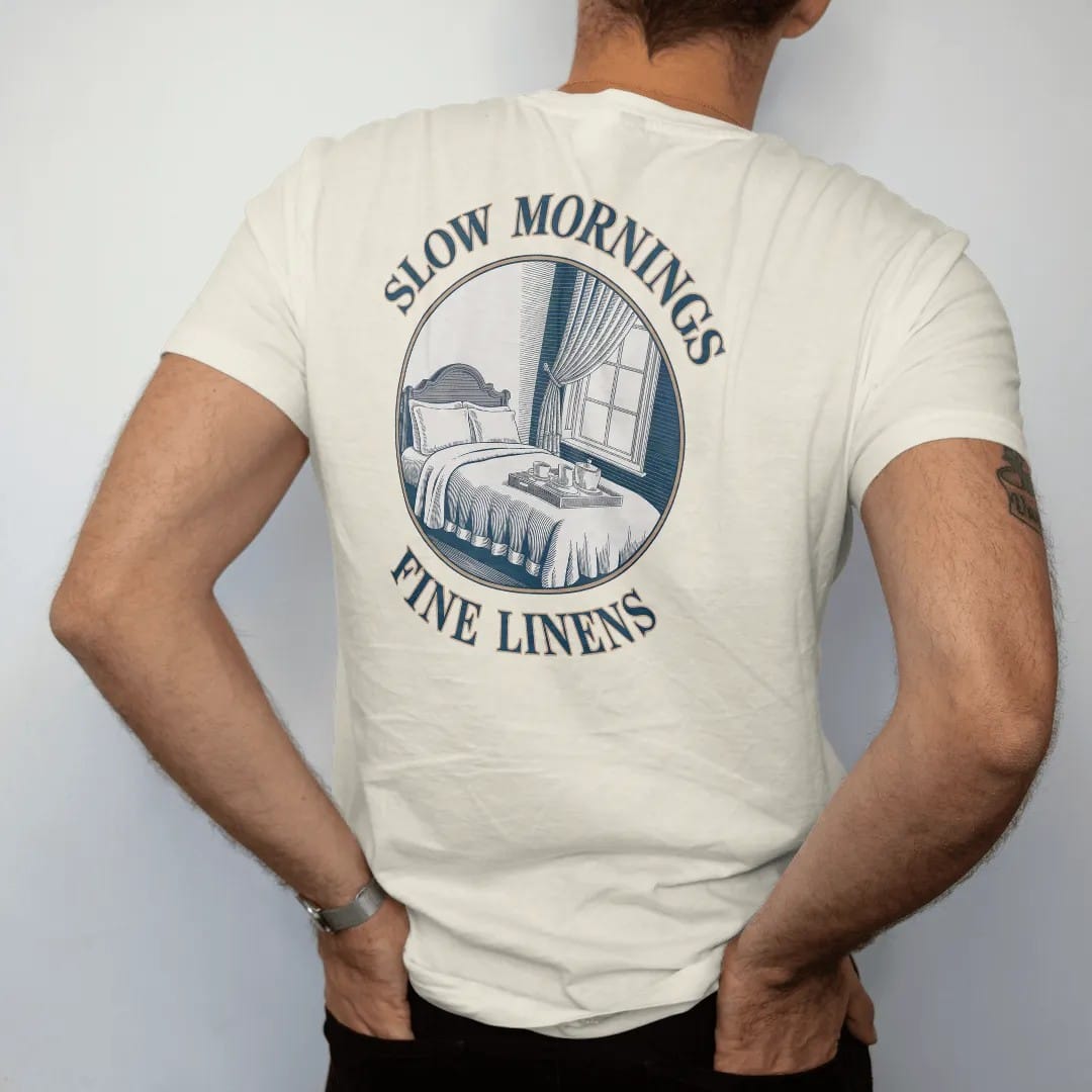 Man wearing a Ivory tee, seen from the back: a made bed with crisp linens, a breakfast tray and an open window with a blowing curtain, under Slow Mornings and Fine Linens, as a fine engraving in navy and antique gold.