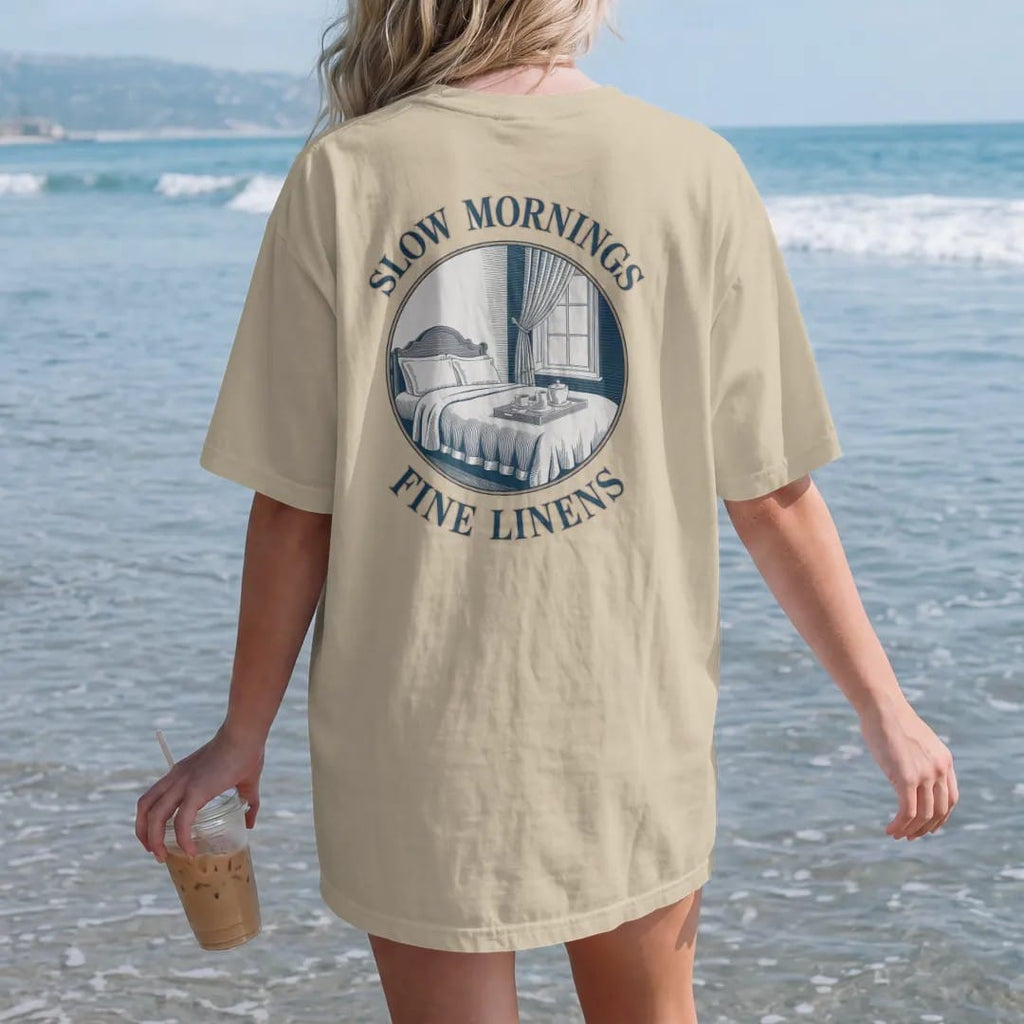 Woman on the beach in a Sandstone tee, back view: a made bed with crisp linens, a breakfast tray and an open window with a blowing curtain, under Slow Mornings and Fine Linens, as a fine engraving in navy and antique gold.