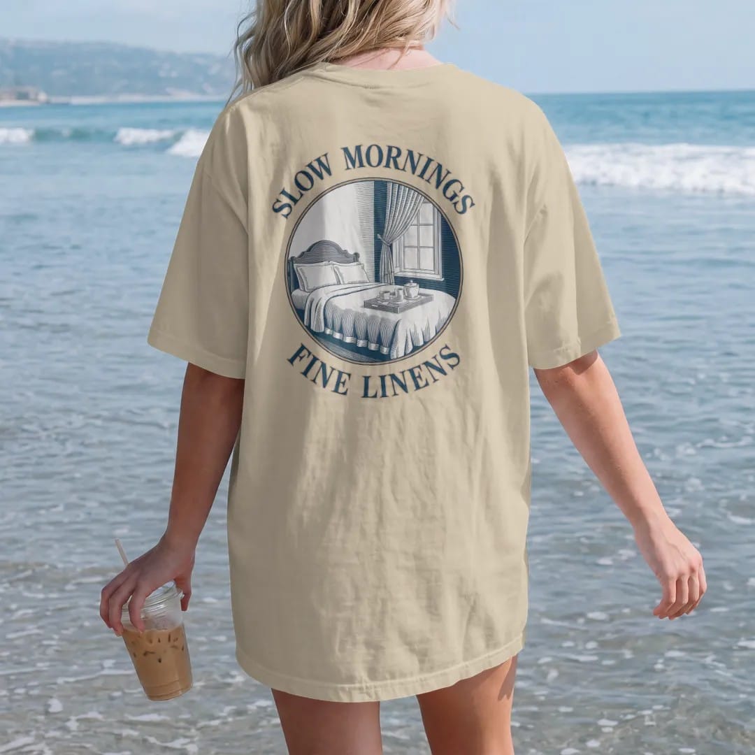 Woman on the beach in a Sandstone tee, back view: a made bed with crisp linens, a breakfast tray and an open window with a blowing curtain, under Slow Mornings and Fine Linens, as a fine engraving in navy and antique gold.