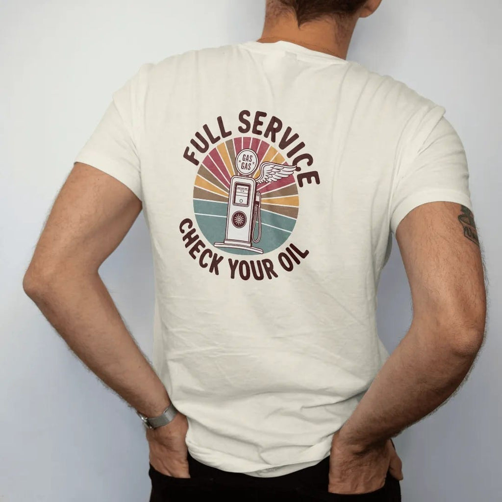 Man wearing a Ivory tee, seen from the back: a round enamel gas-station sign with a vintage pump and winged wheel, with "FULL SERVICE" and "CHECK YOUR OIL" in chunky 70s lettering and small 70s caps, printed in faded, cracked brown, rust red,…