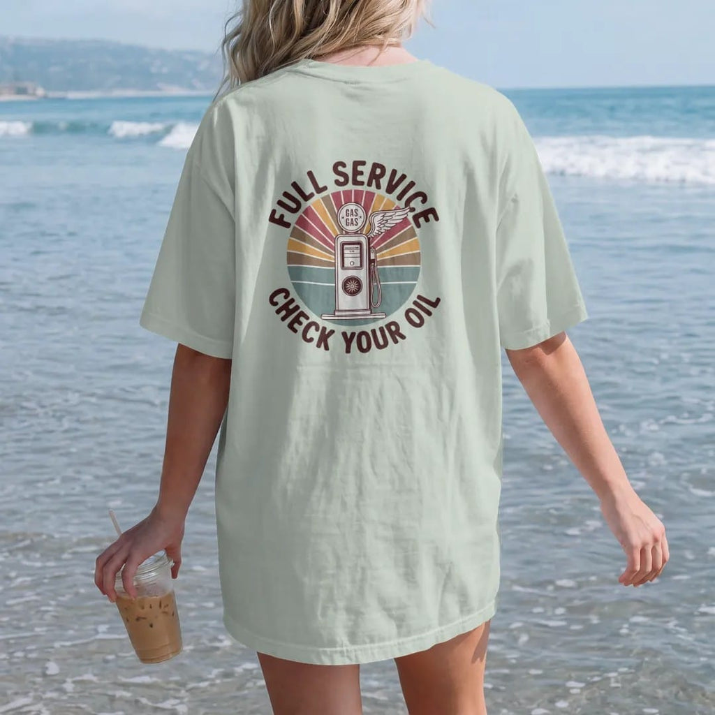 Woman on the beach in a Bay tee, back view: a round enamel gas-station sign with a vintage pump and winged wheel, with "FULL SERVICE" and "CHECK YOUR OIL" in chunky 70s lettering and small 70s caps, printed in faded, cracked brown, rust red,…