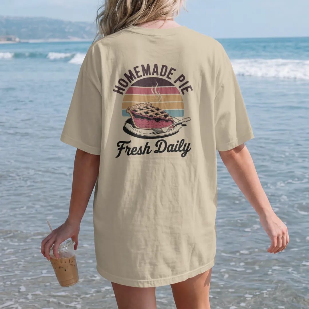 Woman on the beach in a Sandstone tee, back view: a steaming slice of lattice-top cherry pie on a diner plate, with "HOMEMADE PIE" and "FRESH DAILY" in chunky 70s lettering and brush script, printed in faded, cracked brown, rust red, mustard and…