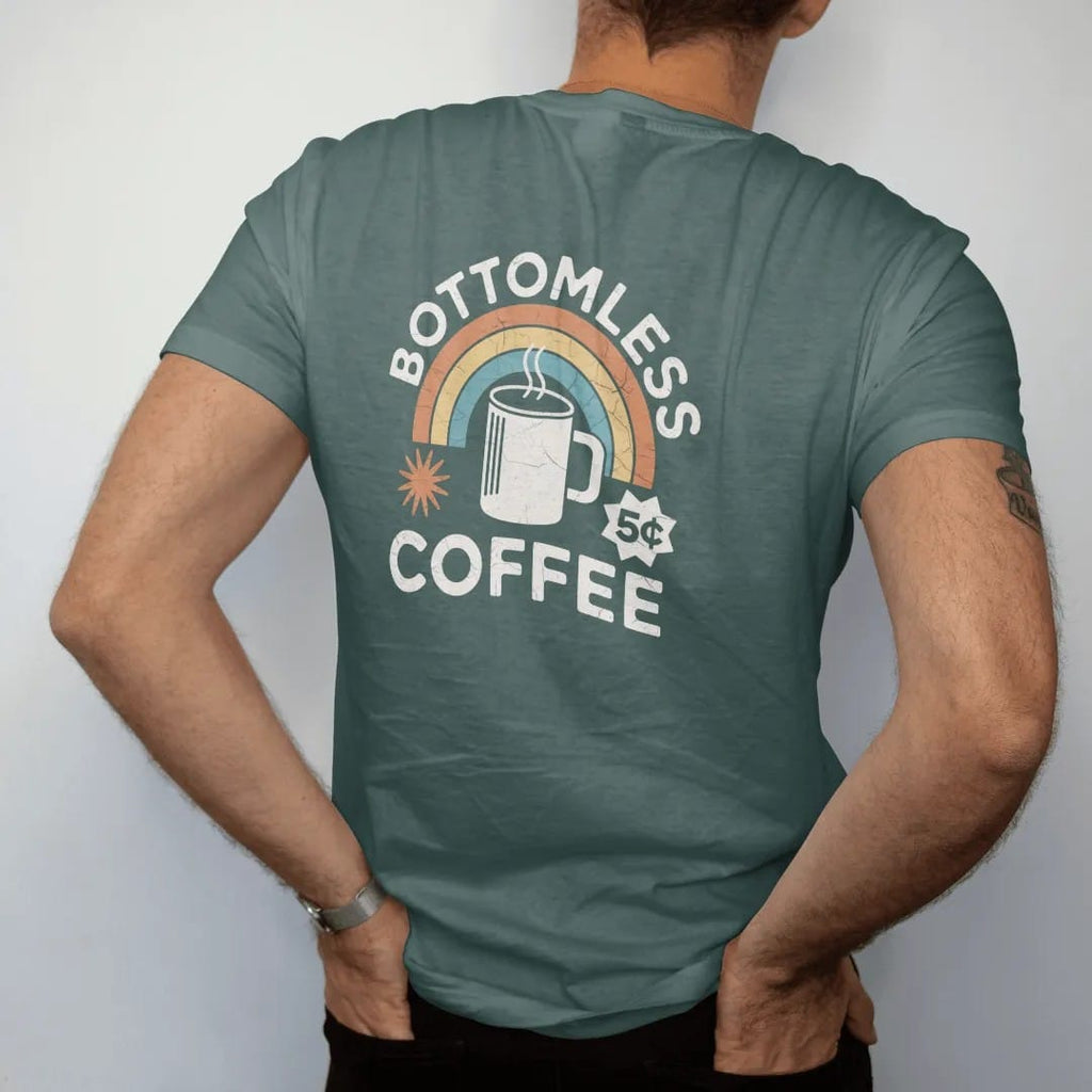 Man wearing a Blue Spruce tee, seen from the back: a thick diner mug with a coffee pot pouring from above, with "BOTTOMLESS COFFEE" and "5¢" in chunky 70s lettering and small 70s caps, printed in faded, cracked cream, mustard, burnt orange and…