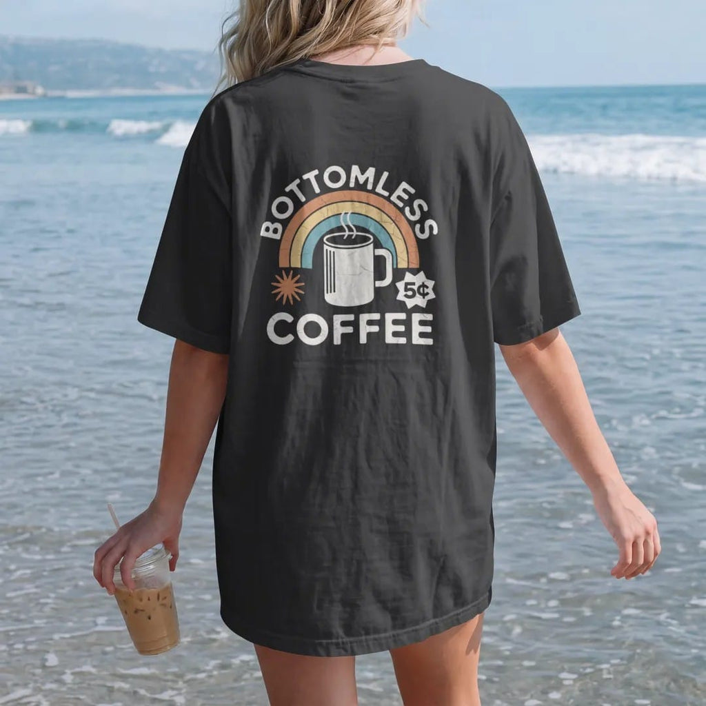 Woman on the beach in a Black tee, back view: a thick diner mug with a coffee pot pouring from above, with "BOTTOMLESS COFFEE" and "5¢" in chunky 70s lettering and small 70s caps, printed in faded, cracked cream, mustard, burnt orange and dusty…