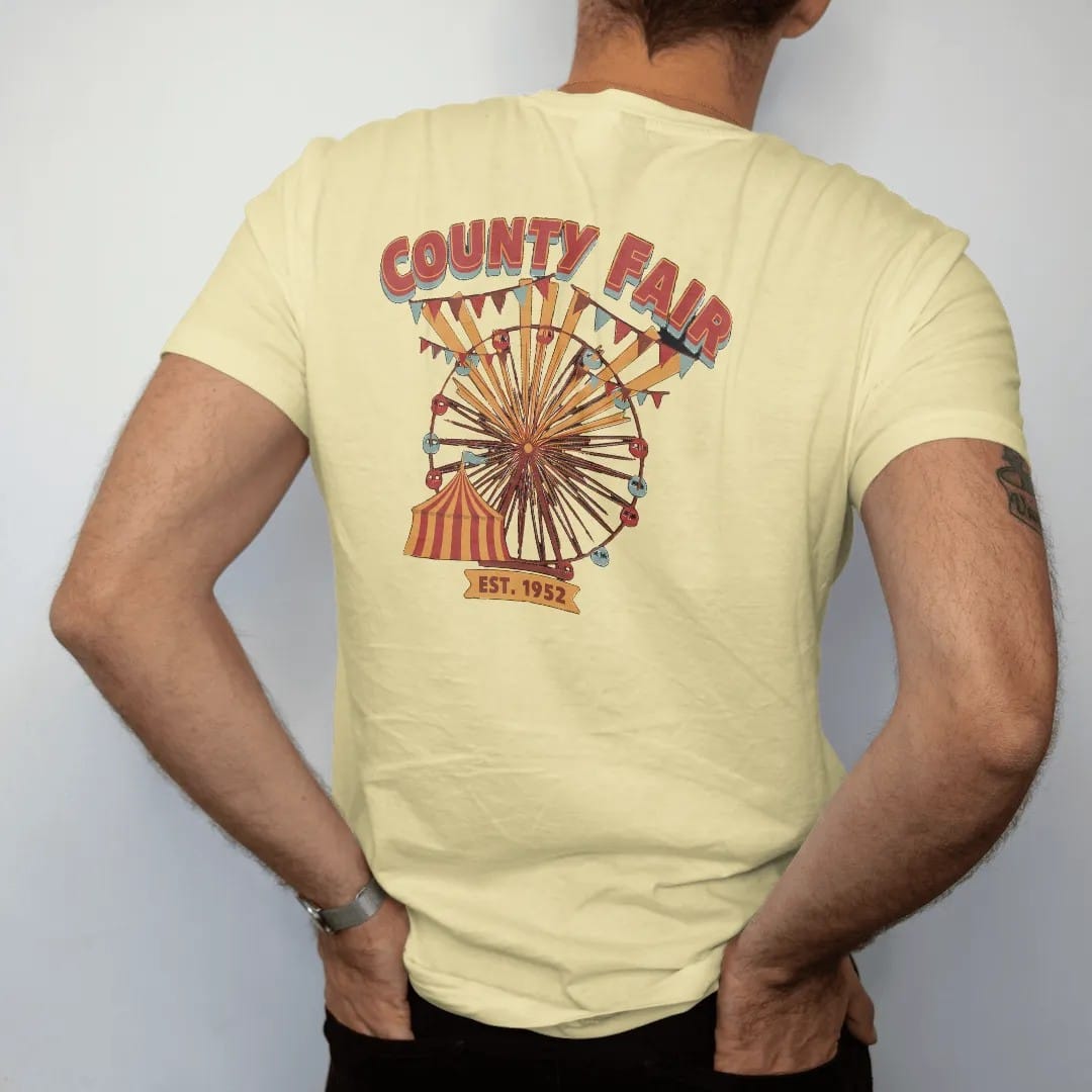 Man wearing a Butter tee, seen from the back: a Ferris wheel and striped fair tent with pennant strings, with "COUNTY FAIR" and "EST. 1952" in chunky 70s lettering and small 70s caps, printed in faded, cracked brown, rust red, mustard and dusty…