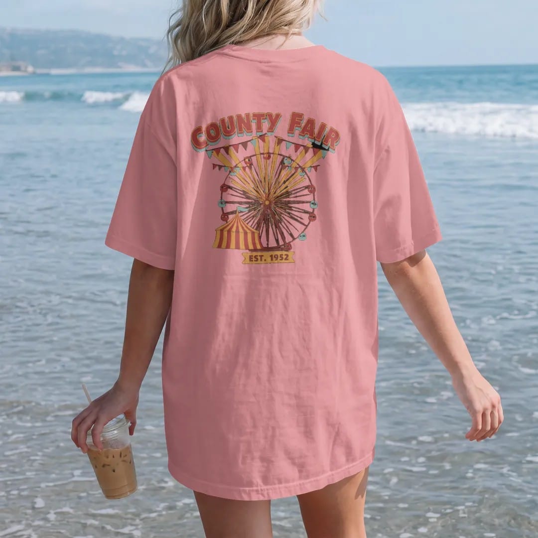 Woman on the beach in a Watermelon tee, back view: a Ferris wheel and striped fair tent with pennant strings, with "COUNTY FAIR" and "EST. 1952" in chunky 70s lettering and small 70s caps, printed in faded, cracked brown, rust red, mustard and…