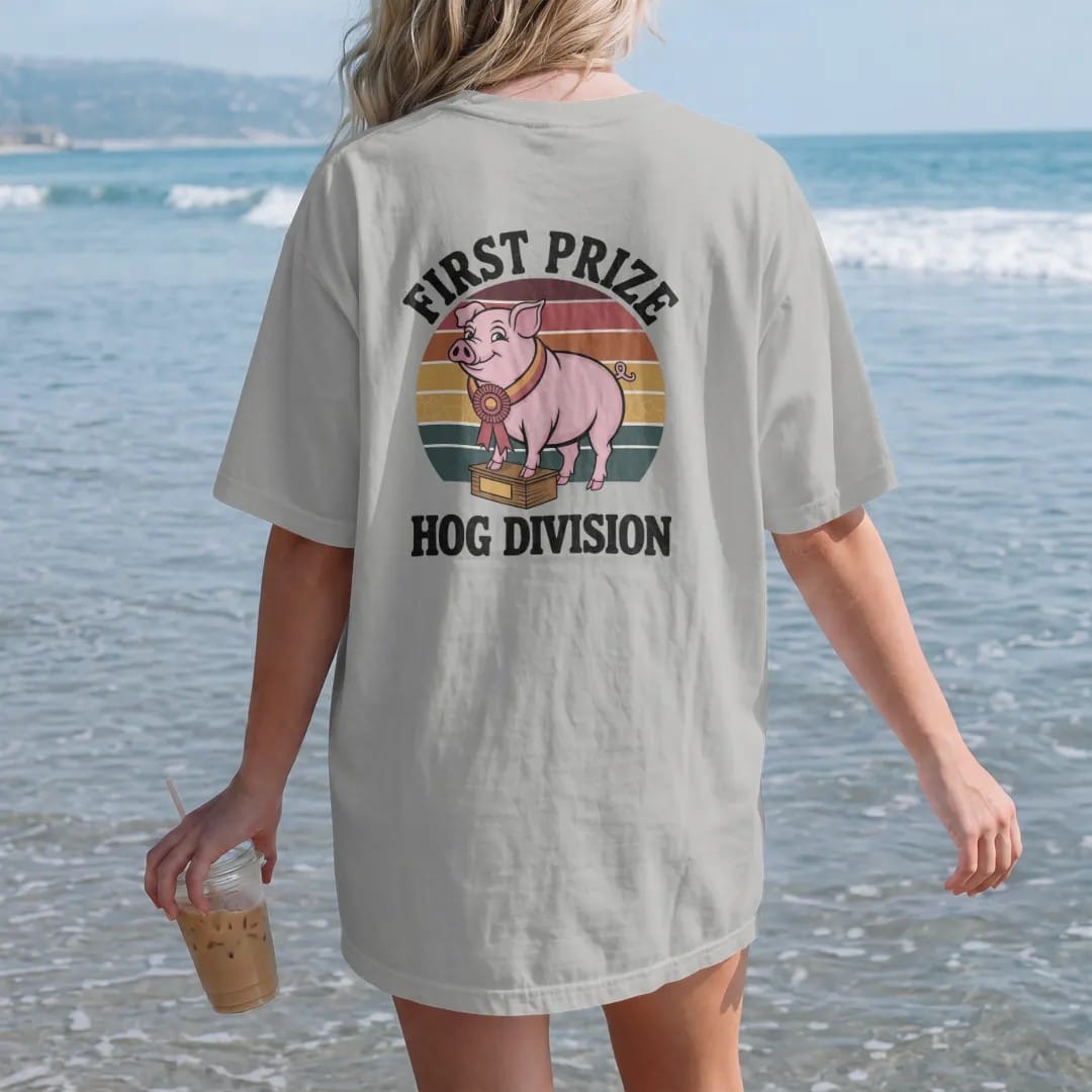 Woman on the beach in a Granite tee, back view: a proud cartoon pig wearing a rosette ribbon on a judging block, with "FIRST PRIZE" and "HOG DIVISION" in chunky 70s lettering and small 70s caps, printed in faded, cracked brown, rust red, mustard…