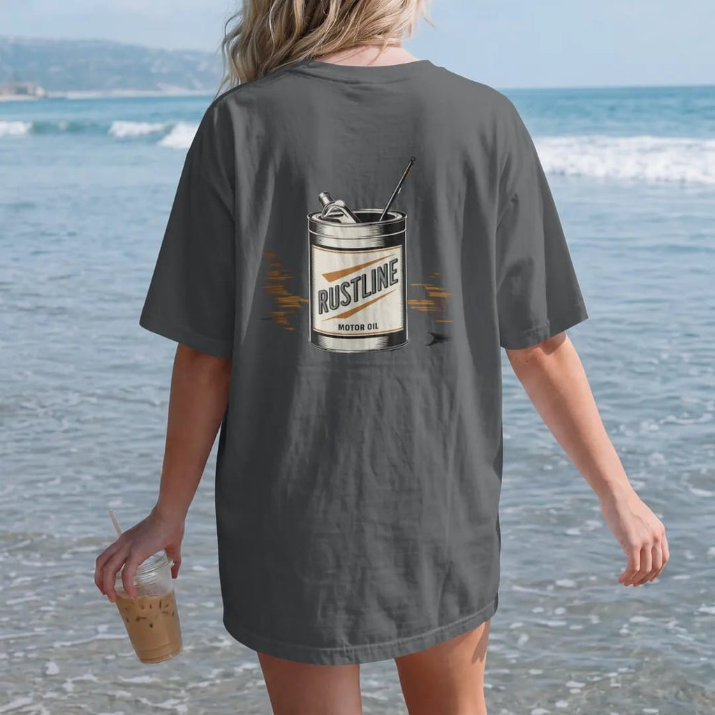 Woman on the beach in a Graphite tee, back view: a dented vintage tin oil can with a racing flag, with "RUSTLINE" and "MOTOR OIL" in chunky 70s lettering and small 70s caps, printed in faded, cracked cream, mustard, burnt orange and dusty teal over…