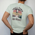 Man wearing a Bay tee, seen from the back: a water tower with a painted heart above a tiny main street, with "SMALL TOWN" and "BIG HEART" in chunky 70s lettering and small 70s caps, printed in faded, cracked brown, rust red, mustard and dusty teal…