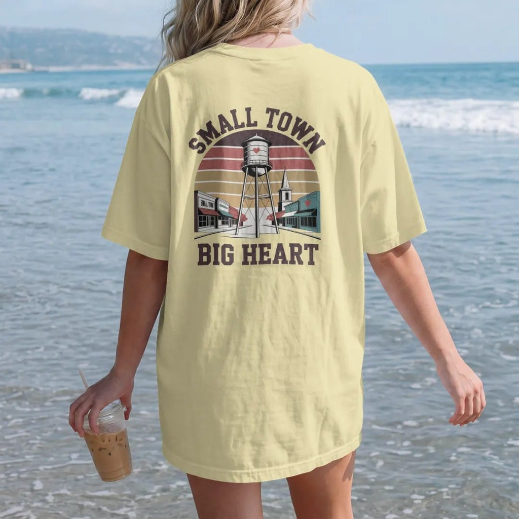 Woman on the beach in a Butter tee, back view: a water tower with a painted heart above a tiny main street, with "SMALL TOWN" and "BIG HEART" in chunky 70s lettering and small 70s caps, printed in faded, cracked brown, rust red, mustard and dusty…