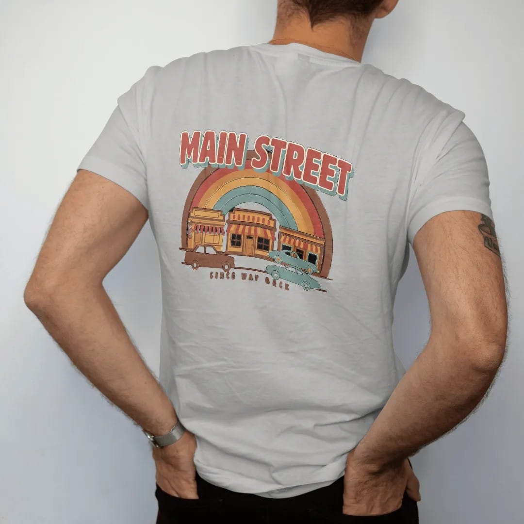 Man wearing a Granite tee, seen from the back: a row of vintage storefronts with striped awnings and old cars, with "MAIN STREET" and "SINCE WAY BACK" in chunky 70s lettering and small 70s caps, printed in faded, cracked brown, rust red, mustard…