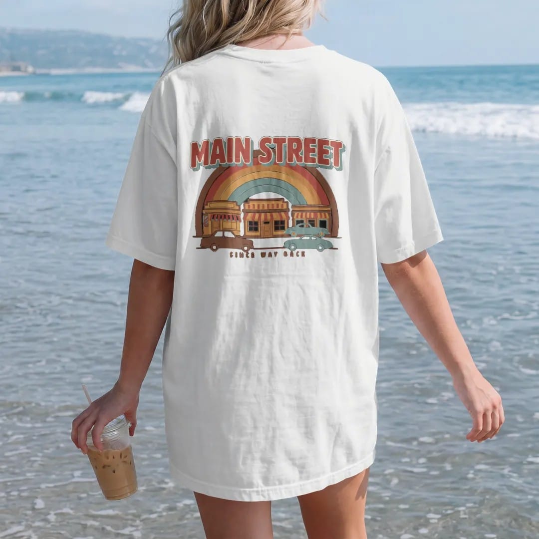 Woman on the beach in a White tee, back view: a row of vintage storefronts with striped awnings and old cars, with "MAIN STREET" and "SINCE WAY BACK" in chunky 70s lettering and small 70s caps, printed in faded, cracked brown, rust red, mustard and…
