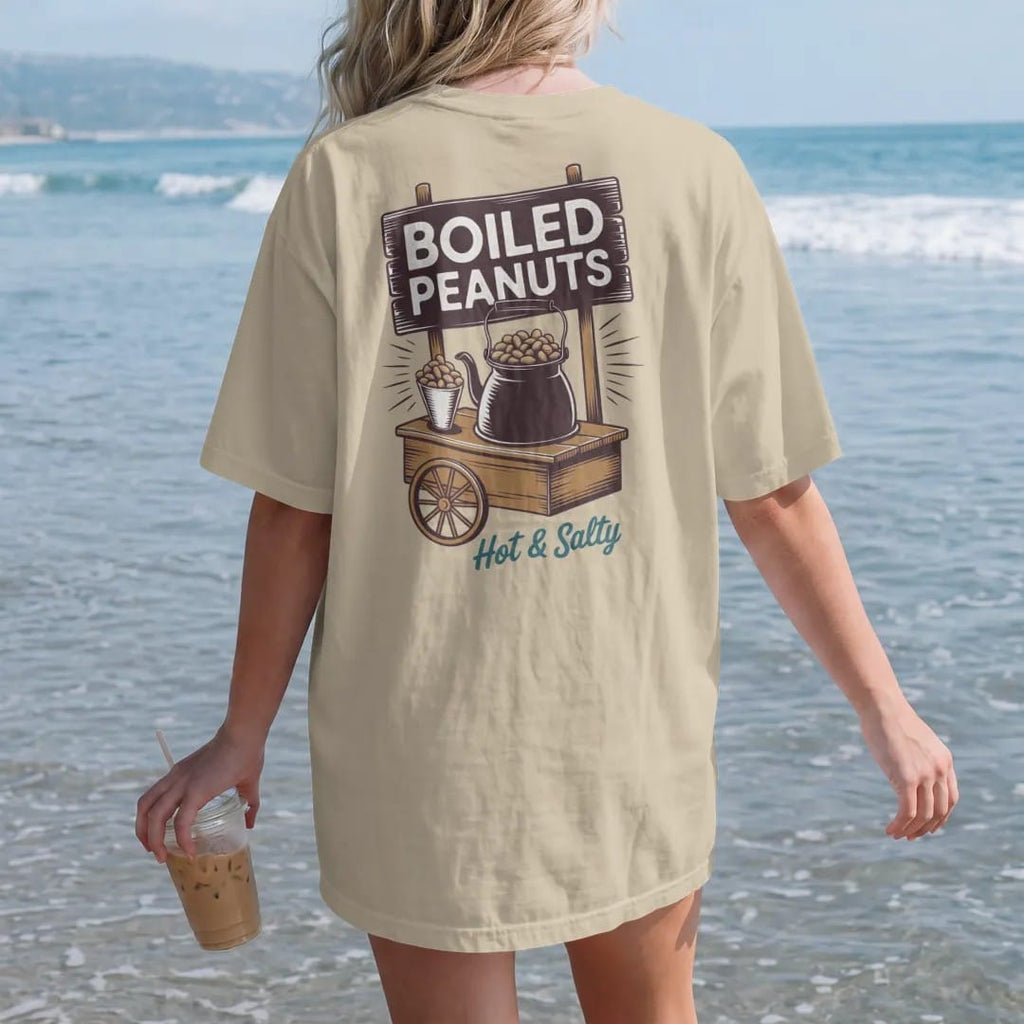Woman on the beach in a Sandstone tee, back view: a steaming kettle of boiled peanuts on a roadside cart, with "BOILED PEANUTS" and "HOT & SALTY" in chunky 70s lettering and brush script, printed in faded, cracked brown, rust red, mustard and dusty…