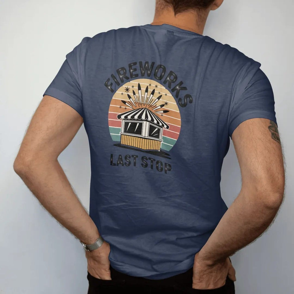 Man wearing a Navy tee, seen from the back: a striped roadside fireworks stand with rockets bursting above, with "FIREWORKS" and "LAST STOP" in chunky 70s lettering and small 70s caps, printed in faded, cracked cream, mustard, burnt orange and…