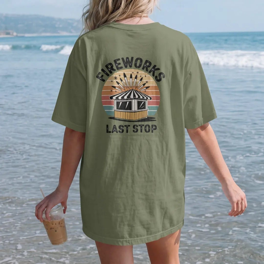 Woman on the beach in a Moss tee, back view: a striped roadside fireworks stand with rockets bursting above, with "FIREWORKS" and "LAST STOP" in chunky 70s lettering and small 70s caps, printed in faded, cracked cream, mustard, burnt orange and…