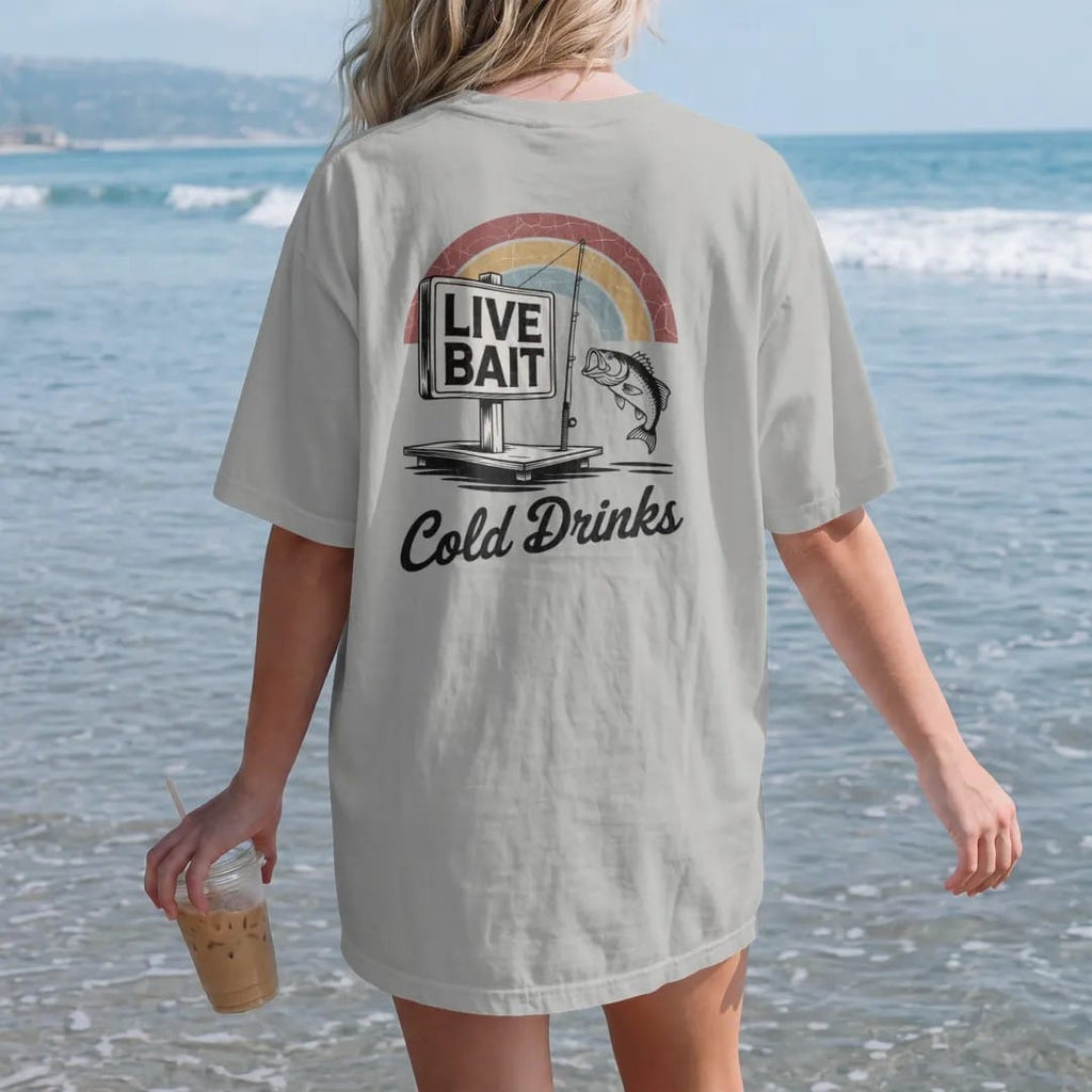 Woman on the beach in a Granite tee, back view: a weathered bait-shop sign with a bobber, fishing pole and jumping bass, with "LIVE BAIT" and "COLD DRINKS" in chunky 70s lettering and brush script, printed in faded, cracked brown, rust red, mustard…