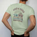 Man wearing a Light Green tee, seen from the back: a seed-packet design with seedlings, a watering can and a farm store, with "FEED & SEED" and "SPRING PLANTING" in chunky 70s lettering and small 70s caps, printed in faded, cracked brown, rust red,…