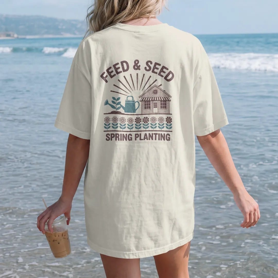 Woman on the beach in a Ivory tee, back view: a seed-packet design with seedlings, a watering can and a farm store, with "FEED & SEED" and "SPRING PLANTING" in chunky 70s lettering and small 70s caps, printed in faded, cracked brown, rust red,…