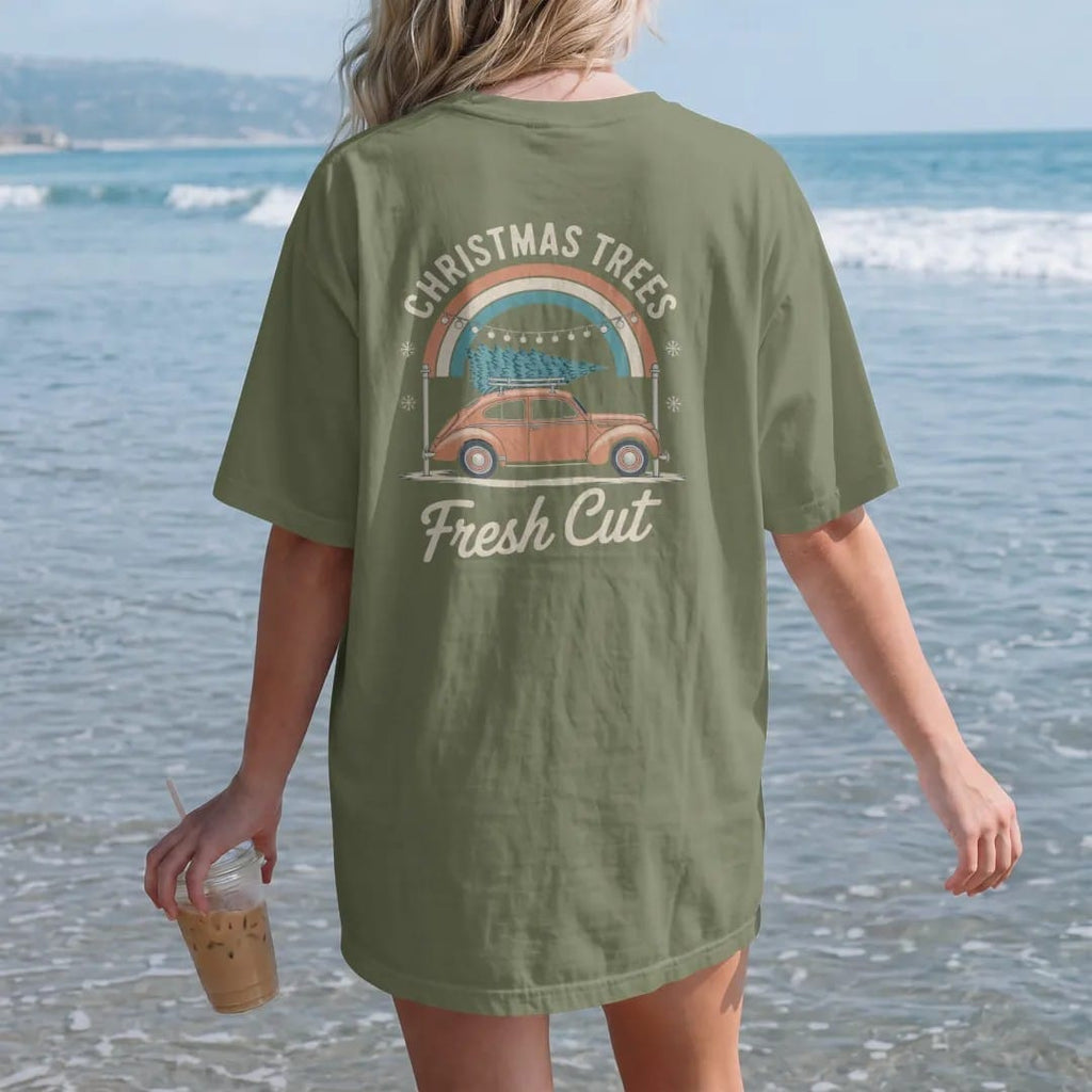 Woman on the beach in a Moss tee, back view: a string-lit tree lot with a pine tied on an old car roof, with "CHRISTMAS TREES" and "FRESH CUT" in chunky 70s lettering and brush script, printed in faded, cracked cream, mustard, burnt orange and…