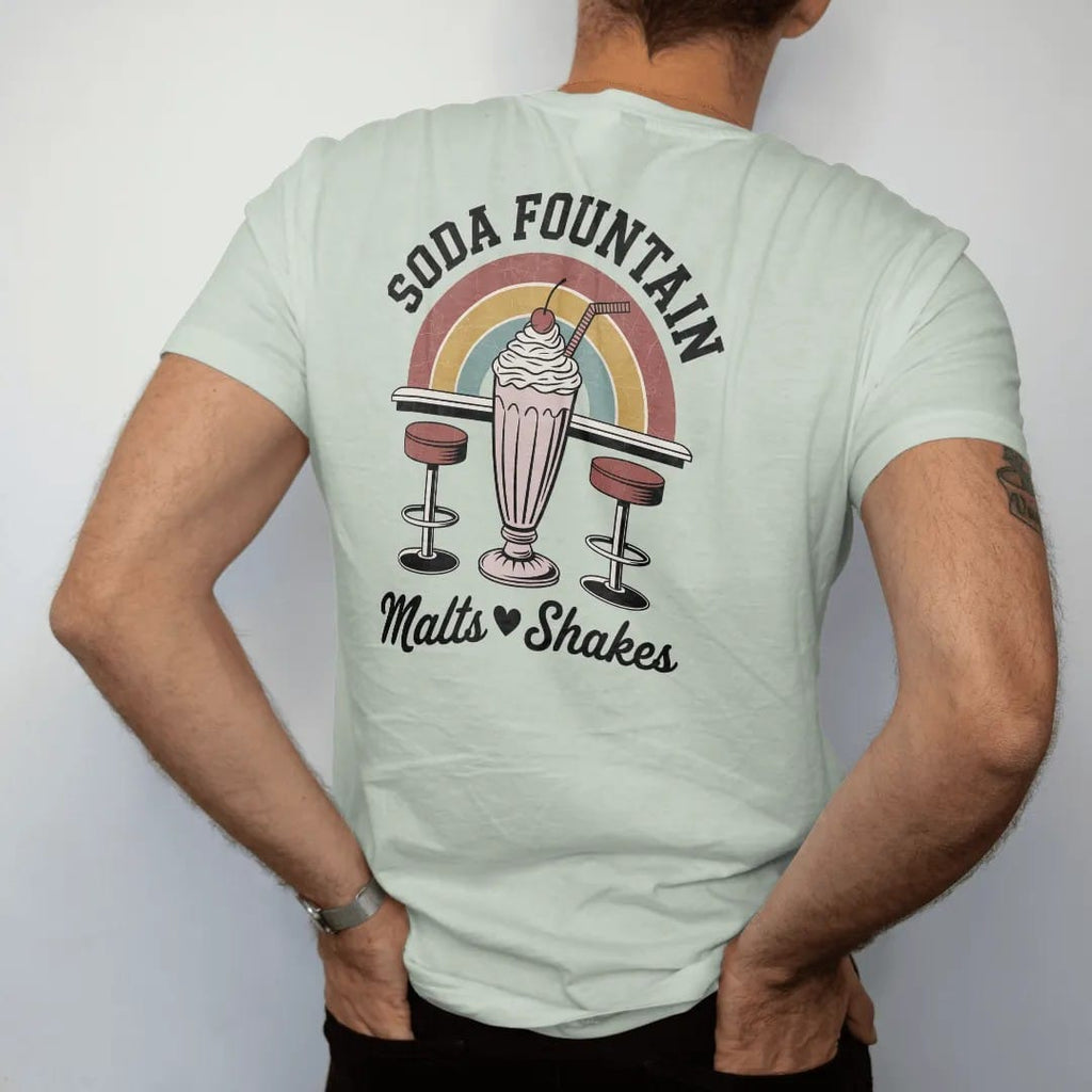 Man wearing a Bay tee, seen from the back: a tall milkshake with cherry and two straws over swivel stools, with "SODA FOUNTAIN" and "MALTS · SHAKES" in chunky 70s lettering and brush script, printed in faded, cracked brown, rust red, mustard and…