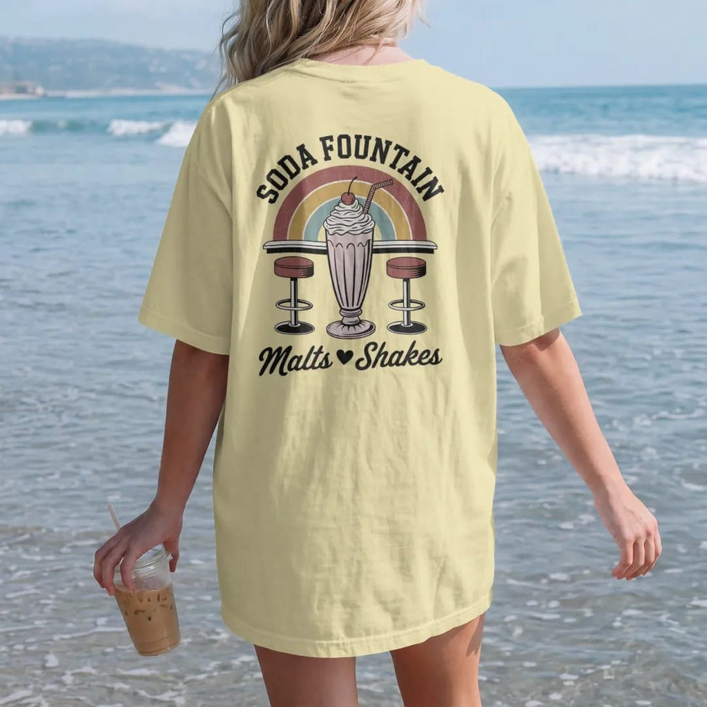 Woman on the beach in a Butter tee, back view: a tall milkshake with cherry and two straws over swivel stools, with "SODA FOUNTAIN" and "MALTS · SHAKES" in chunky 70s lettering and brush script, printed in faded, cracked brown, rust red, mustard…