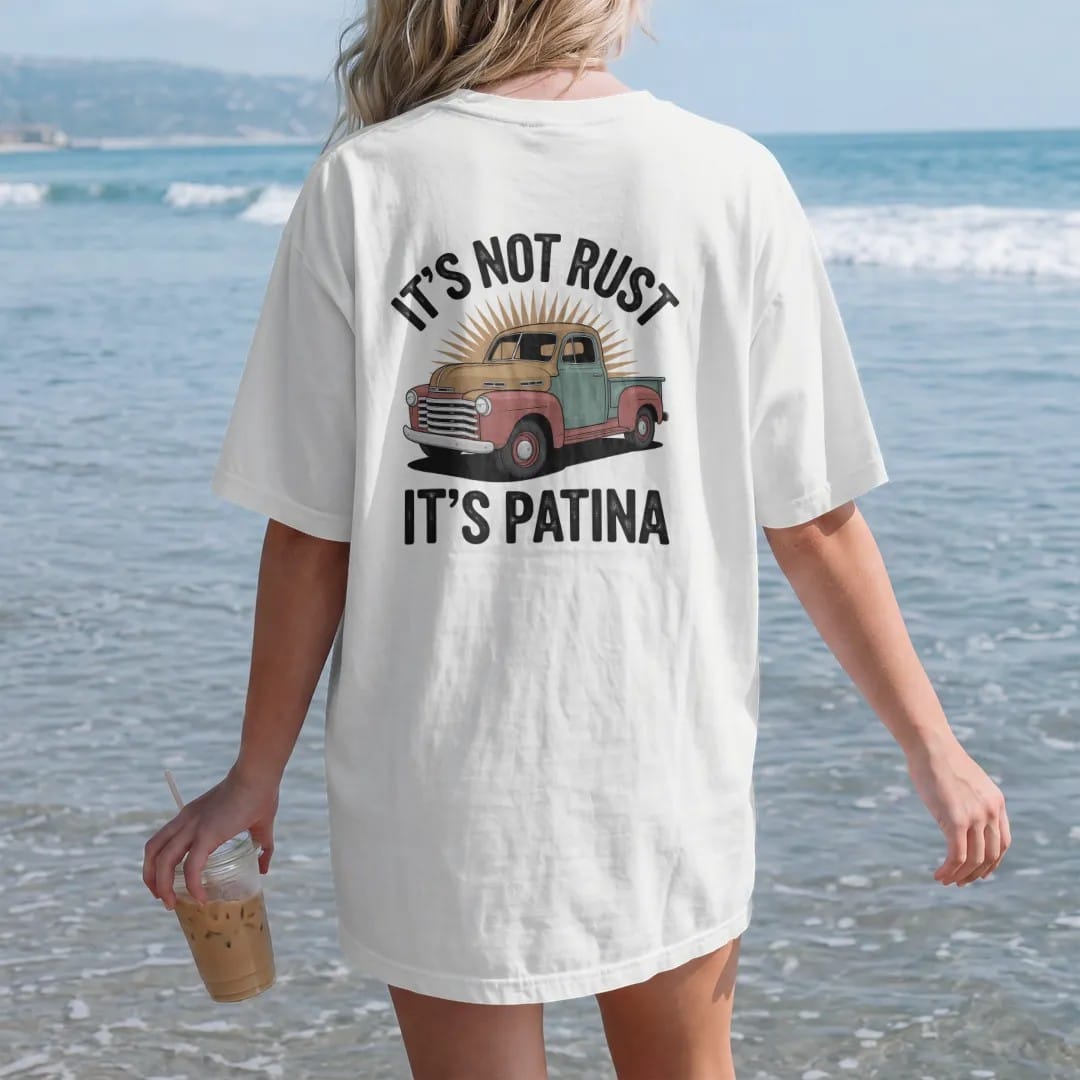 Woman on the beach in a White tee, back view: an old rounded pickup with rusty fenders and faded paint, with "IT'S NOT RUST" and "IT'S PATINA" in chunky 70s lettering and small 70s caps, printed in faded, cracked brown, rust red, mustard and dusty…