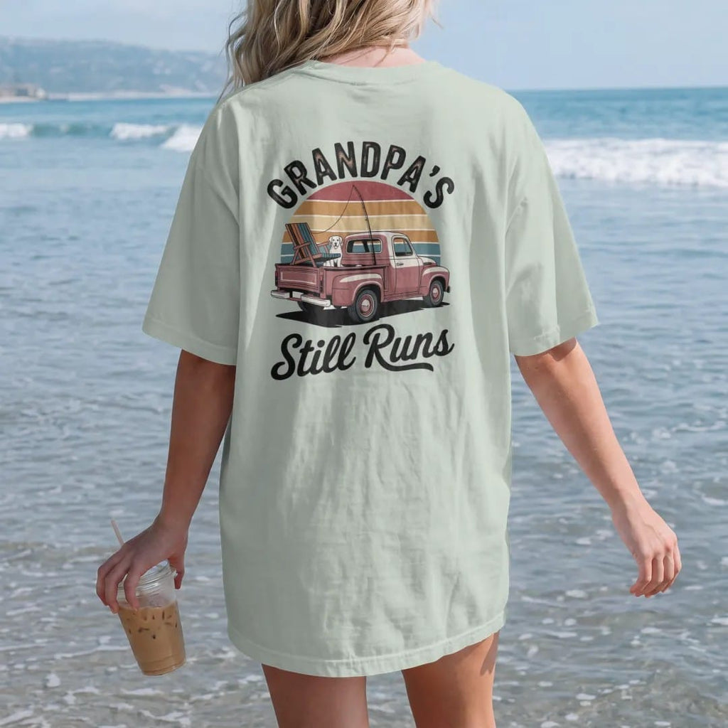 Woman on the beach in a Bay tee, back view: a vintage pickup with a dog, fishing rod and lawn chair, with "GRANDPA'S TRUCK" and "STILL RUNS" in chunky 70s lettering and brush script, printed in faded, cracked brown, rust red, mustard and dusty teal…