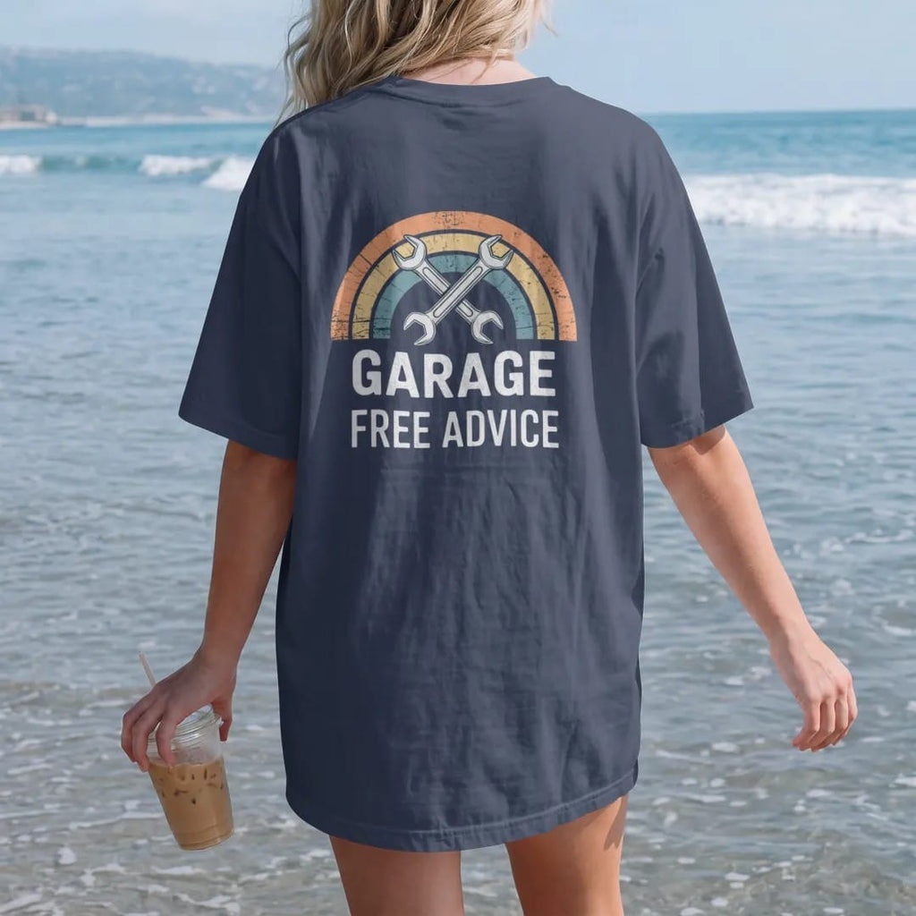 Woman on the beach in a Navy tee, back view: an enamel garage badge with crossed wrenches and a car lift, with "DAD'S GARAGE" and "FREE ADVICE" in chunky 70s lettering and small 70s caps, printed in faded, cracked cream, mustard, burnt orange and…