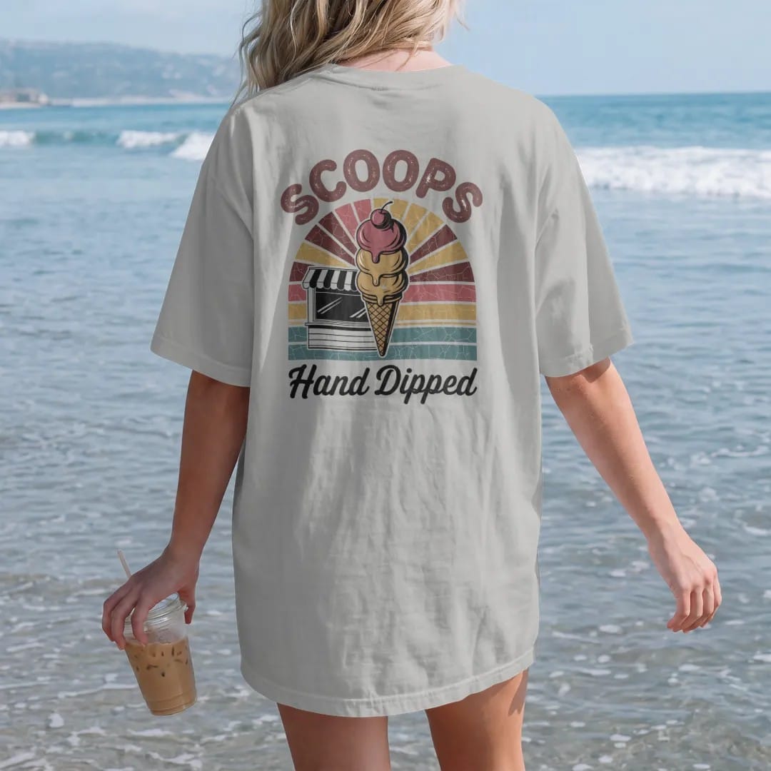 Woman on the beach in a Granite tee, back view: a triple-scoop cone in front of a walk-up ice cream stand, with "SCOOPS" and "HAND DIPPED" in chunky 70s lettering and brush script, printed in faded, cracked brown, rust red, mustard and dusty teal…