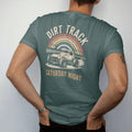 Man wearing a Blue Spruce tee, seen from the back: a vintage sprint car sliding through dust under grandstand lights, with "DIRT TRACK" and "SATURDAY NIGHT" in chunky 70s lettering and small 70s caps, printed in faded, cracked cream, mustard, burnt…