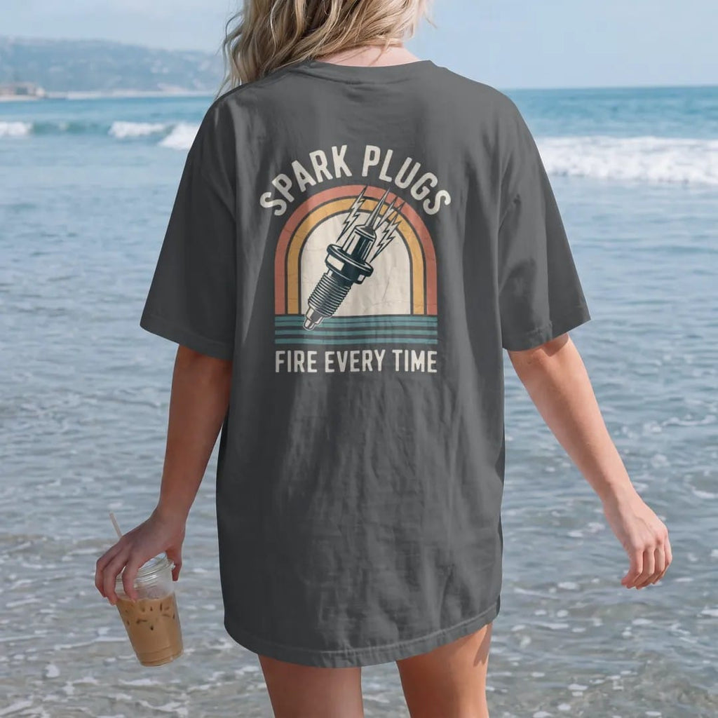 Woman on the beach in a Graphite tee, back view: an oversized vintage spark plug crackling with lightning, with "SPARK PLUGS" and "FIRE EVERY TIME" in chunky 70s lettering and small 70s caps, printed in faded, cracked cream, mustard, burnt orange…