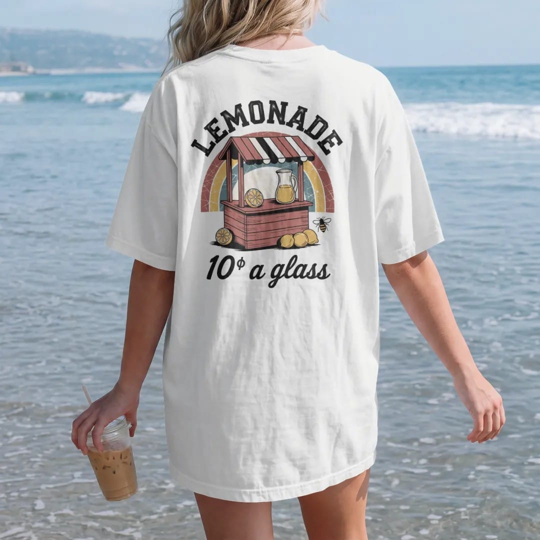 Woman on the beach in a White tee, back view: a wooden lemonade stand with a striped sign, pitcher and lemons, with "LEMONADE" and "10¢ A GLASS" in chunky 70s lettering and brush script, printed in faded, cracked brown, rust red, mustard and dusty…