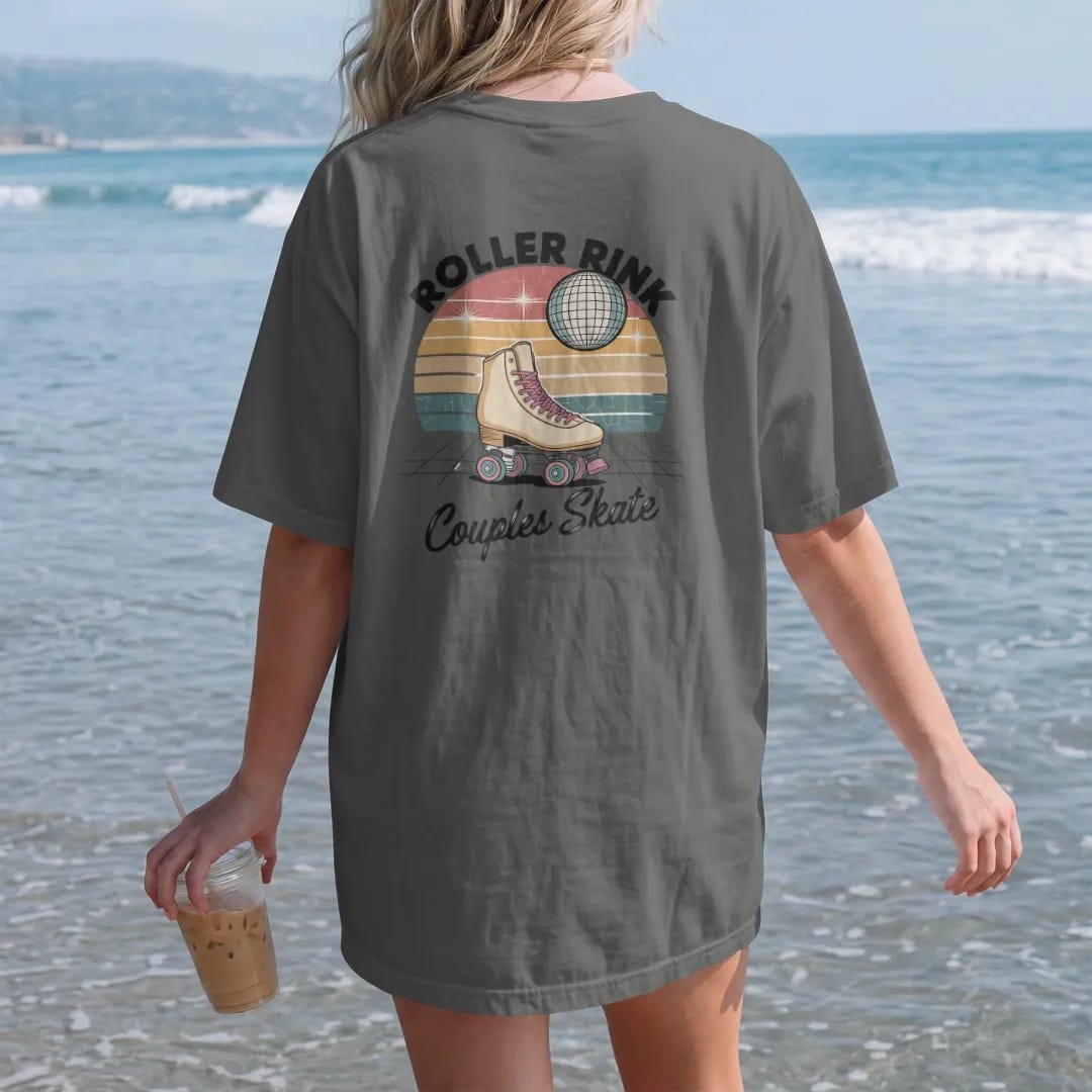 Woman on the beach in a Pepper tee, back view: a vintage roller skate with rainbow wheels under disco sparkles, with "ROLLER RINK" and "COUPLES SKATE" in chunky 70s lettering and brush script, printed in faded, cracked cream, mustard, burnt orange…