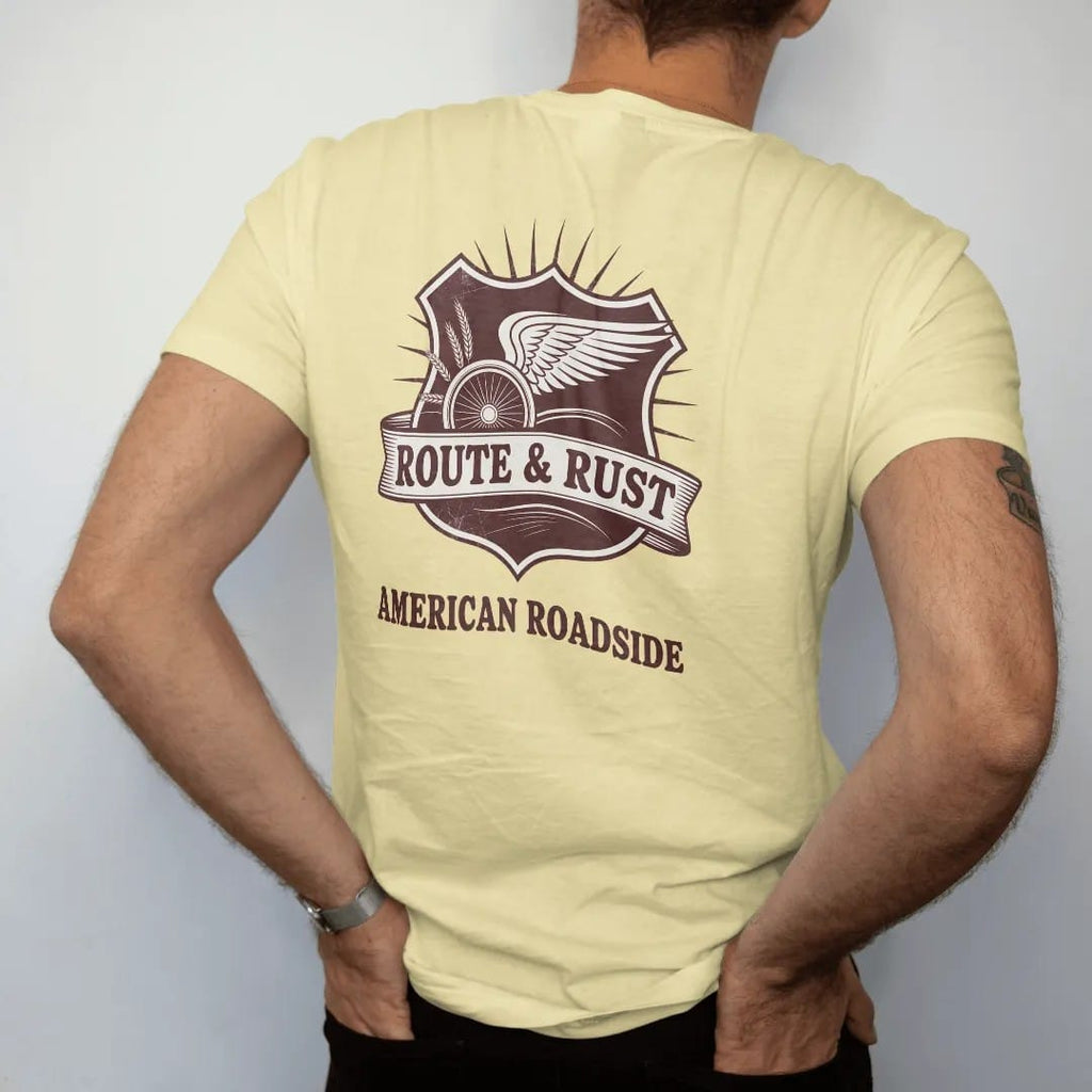 Man wearing a Butter tee, seen from the back: a highway-shield style emblem with a winged wheel and wheat sprigs, with "ROUTE & RUST" and "AMERICAN ROADSIDE" in chunky 70s lettering and small 70s caps, printed in faded, cracked brown, rust red,…