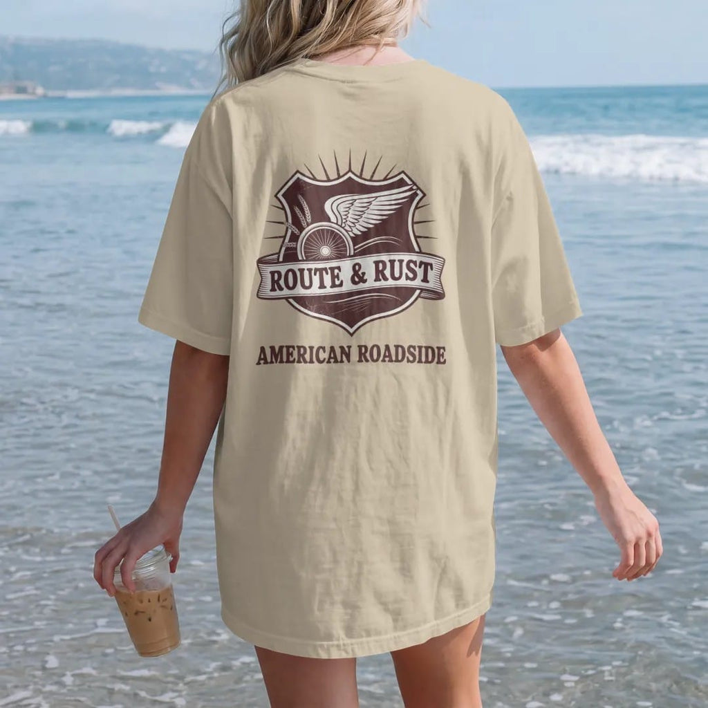 Woman on the beach in a Sandstone tee, back view: a highway-shield style emblem with a winged wheel and wheat sprigs, with "ROUTE & RUST" and "AMERICAN ROADSIDE" in chunky 70s lettering and small 70s caps, printed in faded, cracked brown, rust red,…