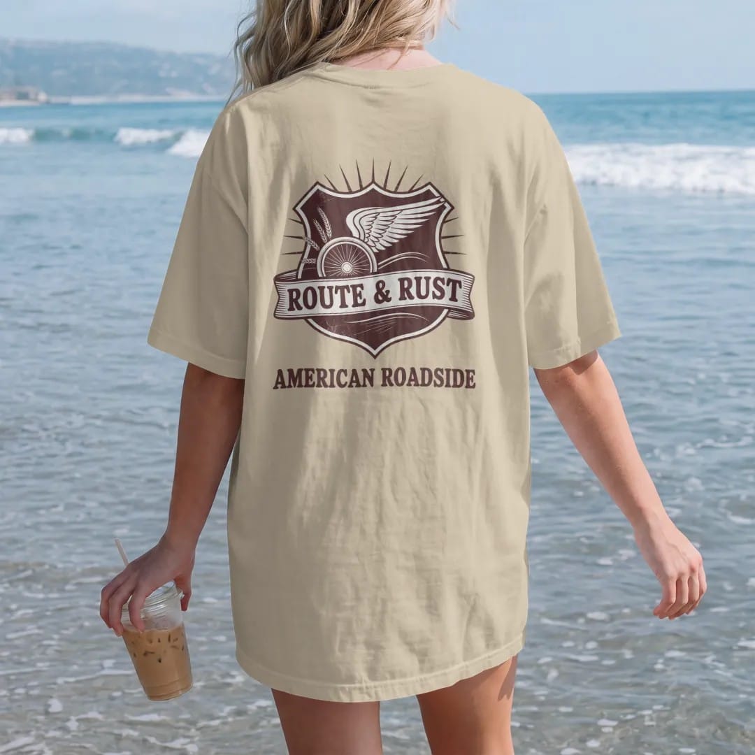 Woman on the beach in a Sandstone tee, back view: a highway-shield style emblem with a winged wheel and wheat sprigs, with "ROUTE & RUST" and "AMERICAN ROADSIDE" in chunky 70s lettering and small 70s caps, printed in faded, cracked brown, rust red,…