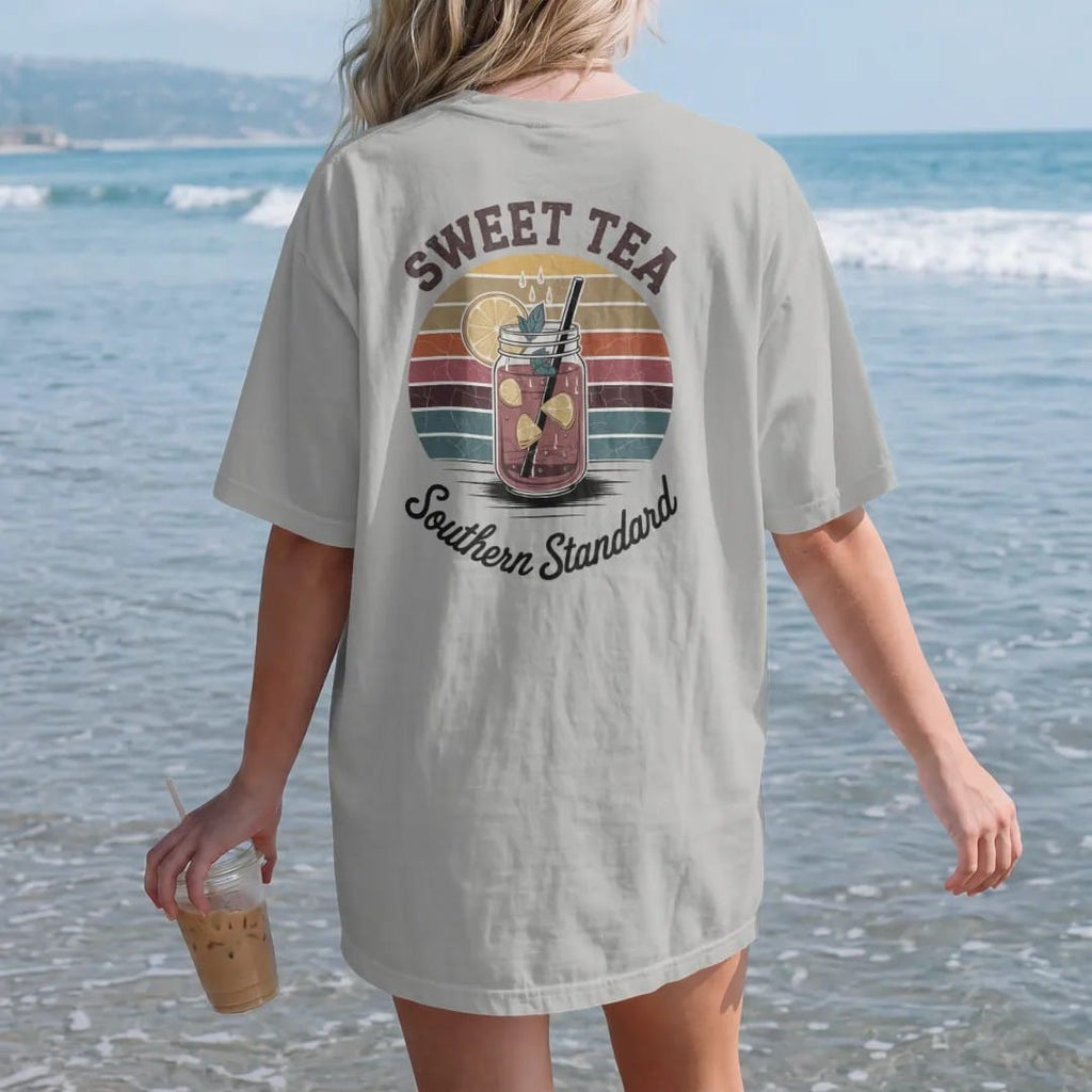 Woman on the beach in a Granite tee, back view: a mason-jar glass of iced tea with lemon and a magnolia blossom, with "SWEET TEA" and "SOUTHERN STANDARD" in chunky 70s lettering and brush script, printed in faded, cracked brown, rust red, mustard…