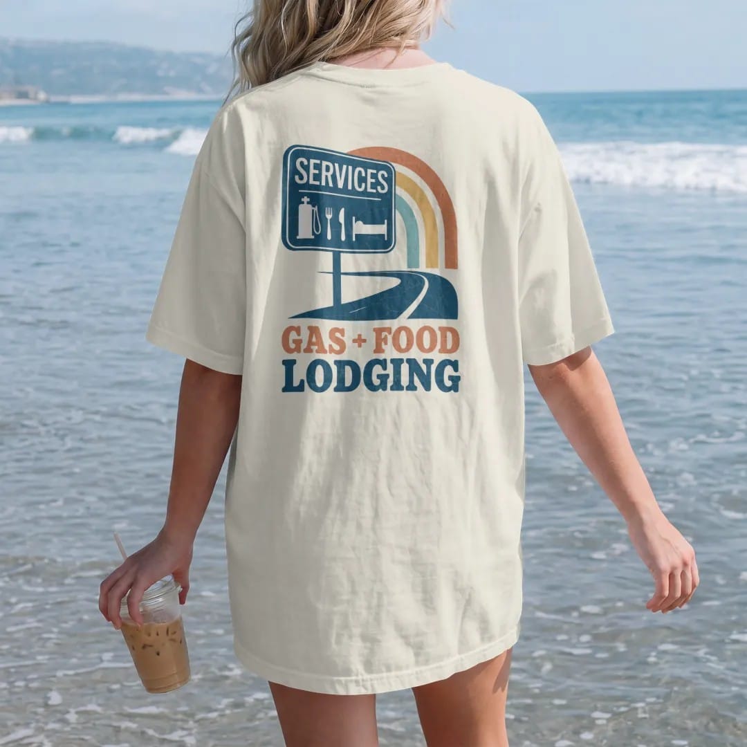 Woman on the beach in a Ivory tee, back view: a highway services sign with pump, fork-and-knife and bed icons at sunset, with "GAS · FOOD · LODGING" in chunky 70s lettering, printed in faded, cracked cream, mustard, burnt orange and dusty teal over…