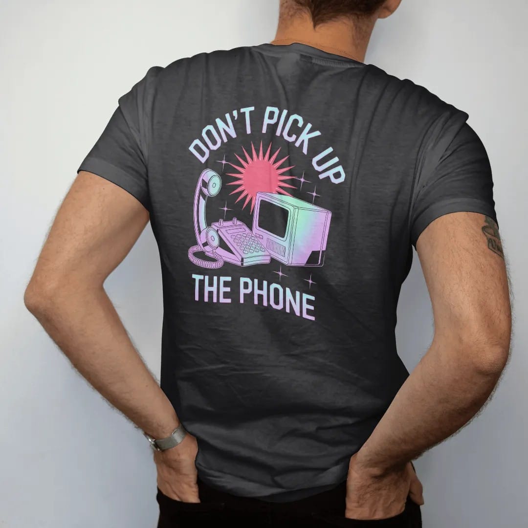 Man wearing a Black tee, seen from the back: a cordless house phone linked by lightning bolts to a boxy computer, with "DON'T PICK UP" and "THE PHONE" in liquid-chrome bubble letters and small pixel type, printed in glossy gunmetal chrome, cobalt,…