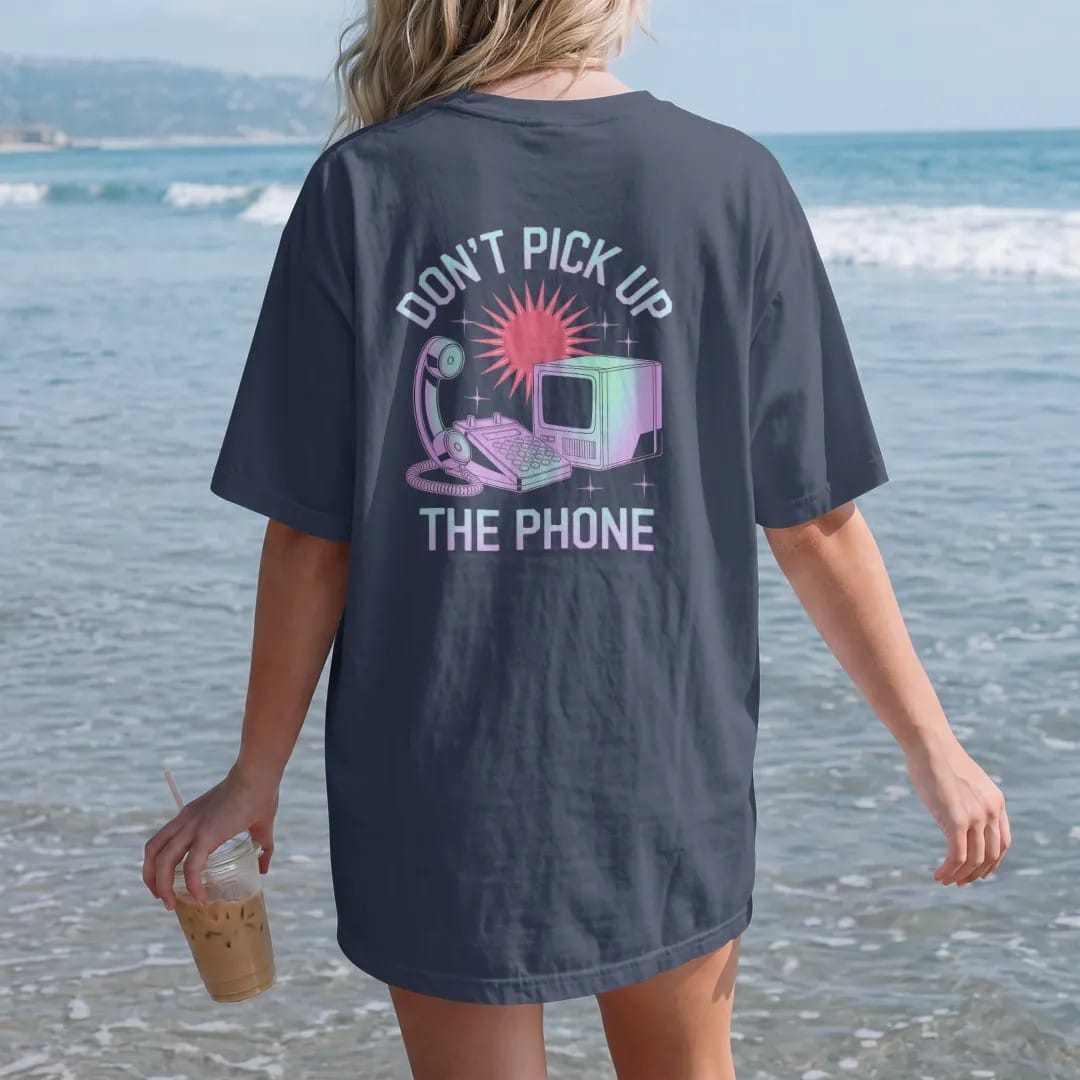 Woman on the beach in a Navy tee, back view: a cordless house phone linked by lightning bolts to a boxy computer, with "DON'T PICK UP" and "THE PHONE" in liquid-chrome bubble letters and small pixel type, printed in glossy gunmetal chrome, cobalt,…