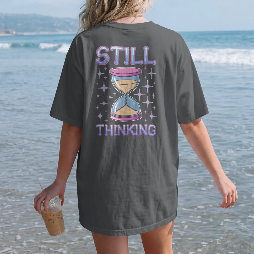 Woman on the beach in a Graphite tee, back view: a giant pixel hourglass cursor with falling sand pixels, with "STILL" and "THINKING" in liquid-chrome bubble letters and small pixel type, printed in glossy chrome silver, baby blue, bubblegum pink…