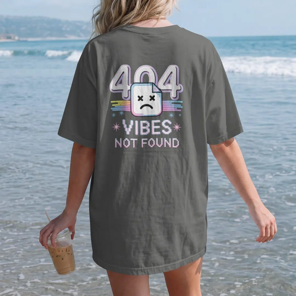 Woman on the beach in a Pepper tee, back view: a broken-page icon with a sad pixel face and glitchy numerals, with "404" and "VIBES NOT FOUND" in liquid-chrome bubble letters and small pixel type, printed in glossy chrome silver, baby blue,…