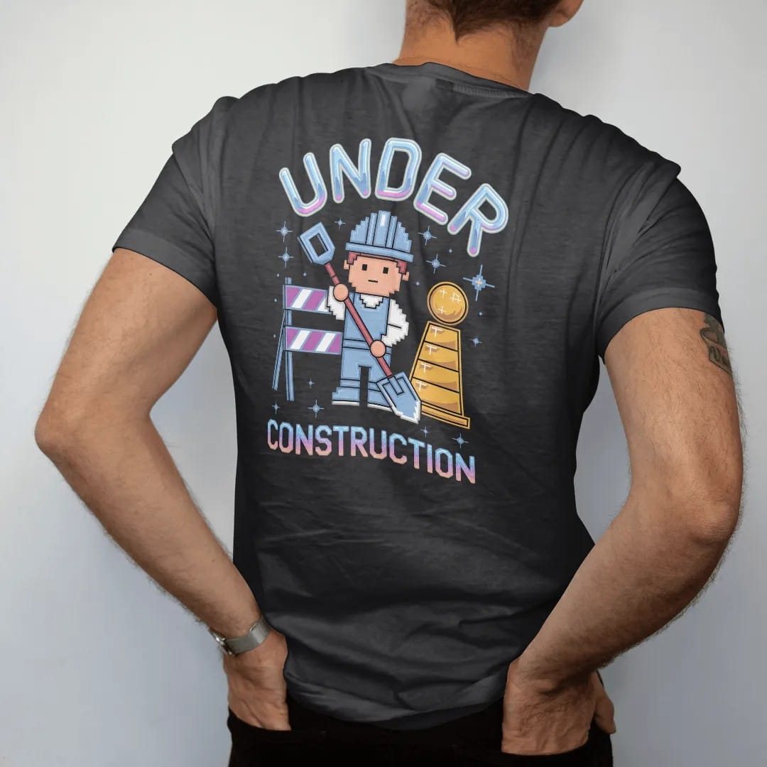 Man wearing a Black tee, seen from the back: a pixel construction worker beside a striped barricade, with "UNDER" and "CONSTRUCTION" in liquid-chrome bubble letters and small pixel type, printed in glossy gunmetal chrome, cobalt, hot magenta and…