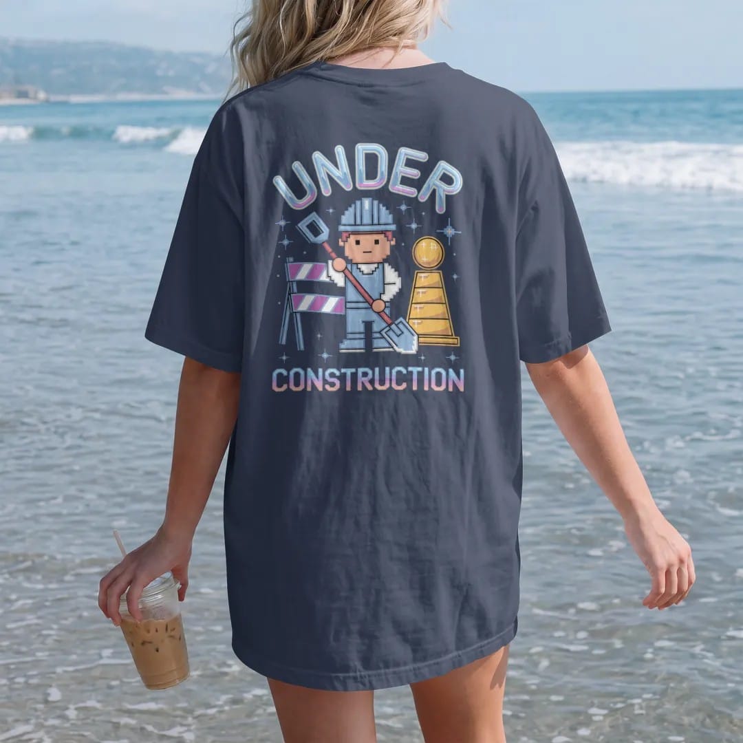 Woman on the beach in a Navy tee, back view: a pixel construction worker beside a striped barricade, with "UNDER" and "CONSTRUCTION" in liquid-chrome bubble letters and small pixel type, printed in glossy gunmetal chrome, cobalt, hot magenta and…