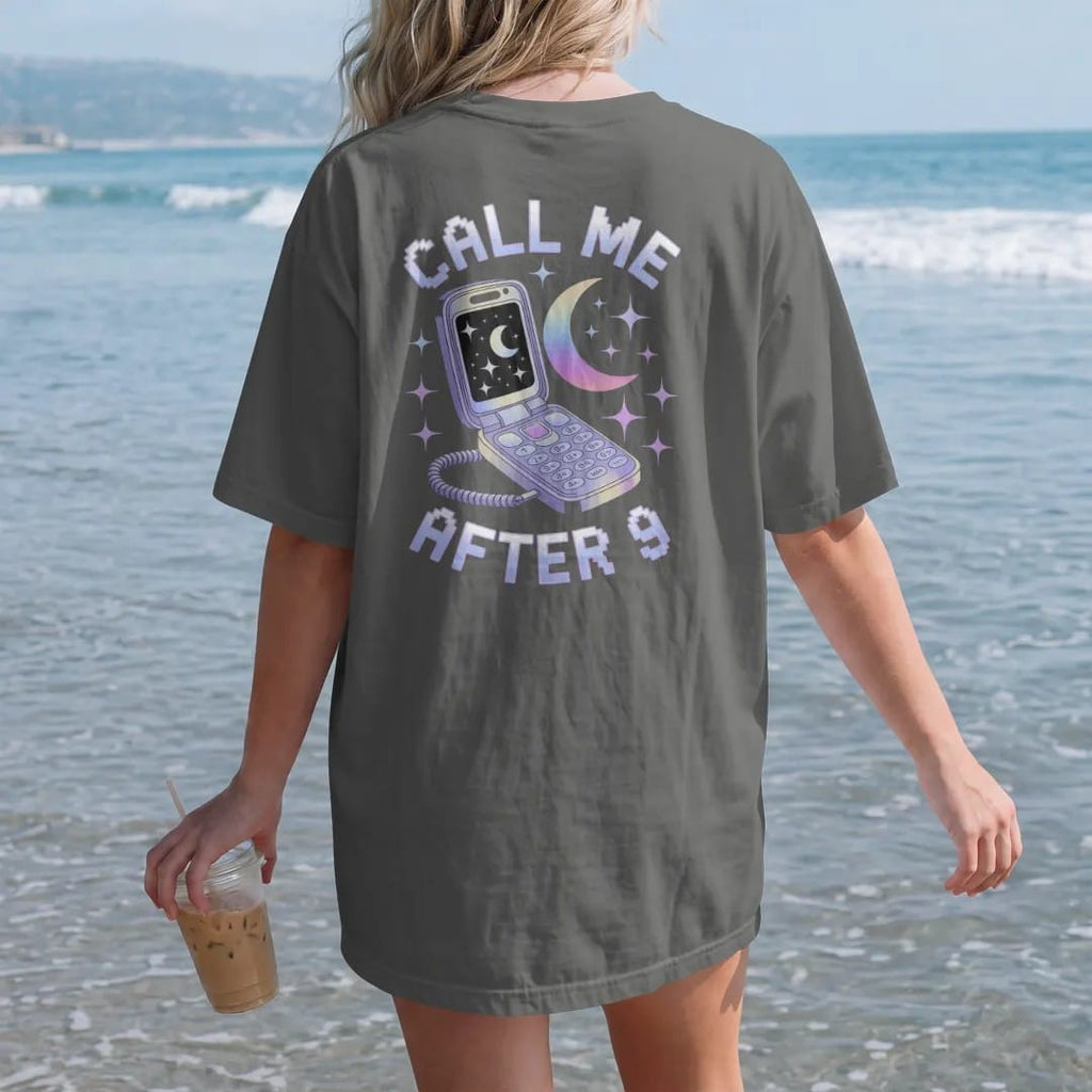 Woman on the beach in a Pepper tee, back view: an open silver flip phone with a moon on its screen, with "CALL ME" and "AFTER 9" in liquid-chrome bubble letters and small pixel type, printed in glossy gunmetal chrome, cobalt, hot magenta and violet…