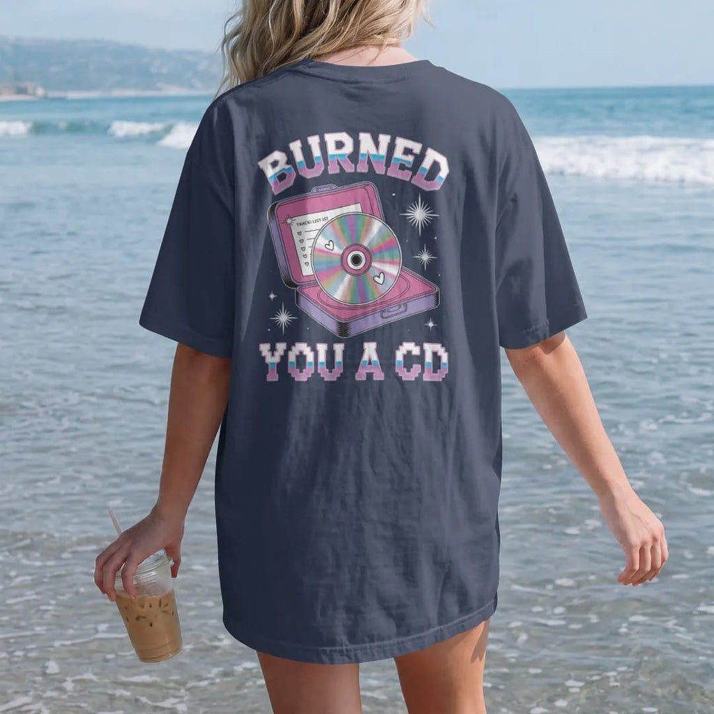 Woman on the beach in a Navy tee, back view: a rainbow-sheen disc in a cracked jewel case with a handwritten list, with "BURNED" and "YOU A CD" in liquid-chrome bubble letters and small pixel type, printed in glossy gunmetal chrome, cobalt, hot…