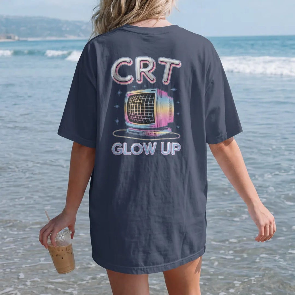Woman on the beach in a Navy tee, back view: a chunky CRT monitor with a glowing scanline screen, with "CRT" and "GLOW UP" in liquid-chrome bubble letters and small pixel type, printed in glossy chrome silver, baby blue, bubblegum pink and violet…