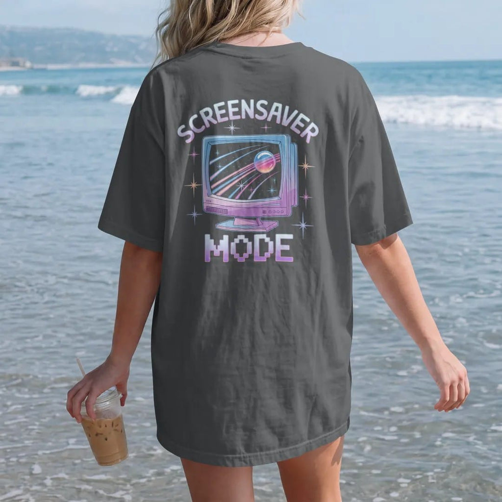 Woman on the beach in a Graphite tee, back view: a CRT monitor showing a starfield screensaver, with "SCREENSAVER" and "MODE" in liquid-chrome bubble letters and small pixel type, printed in glossy chrome silver, baby blue, bubblegum pink and…